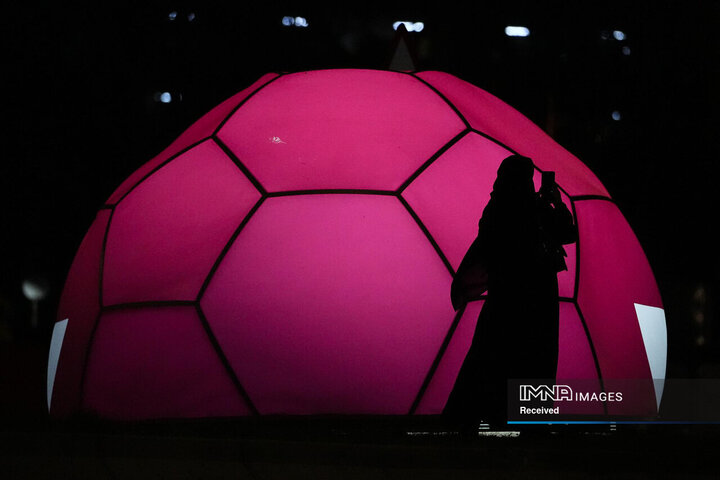 زنی با لباس سنتی قطری از تلفن همراه برای ثبت تصاویر در مقابل توپ فوتبالی که به رنگ پرچم قطر روشن شده است استفاده می کند.